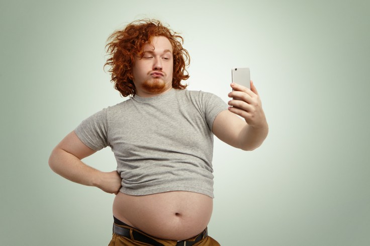 Funny overweight plump man with duck lips wearing undersize t-shirt with belly hanging out of pants, keeping hand on waist, posing for selfie, holding cell phone, trying to seem attractive and sexy